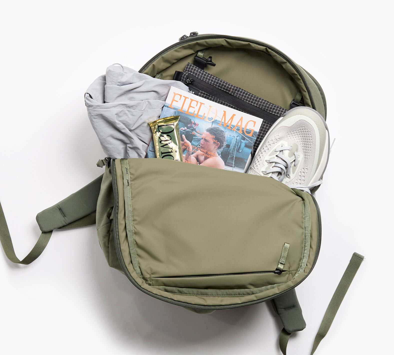 Olive-green backpack open showing white sneakers, a gray t-shirt, and a magazine cover reading 'FIELD MAG'.
