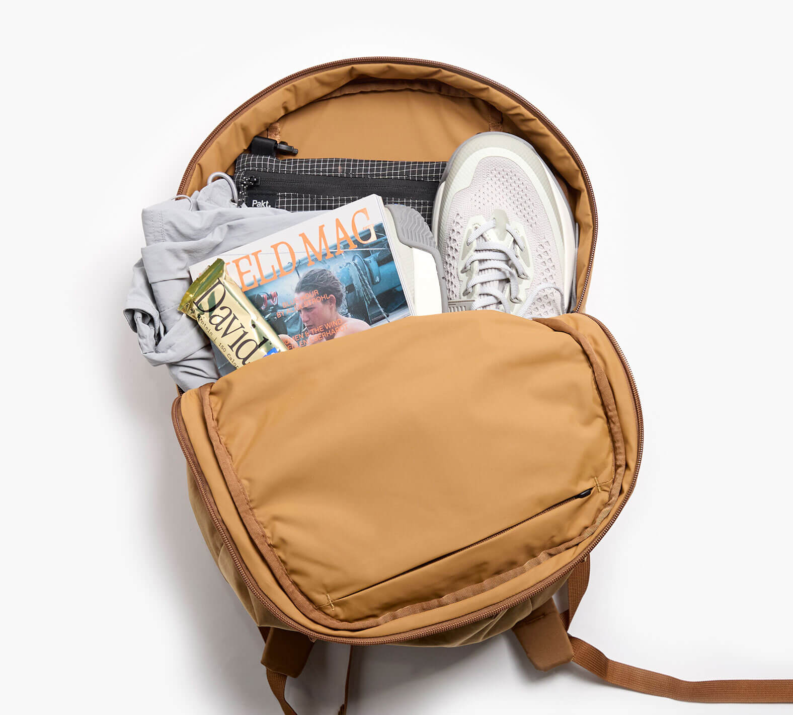Tan MODE backpack open with sneakers, folded shirt, 'ELD MAG' magazine, 'David' snack wrapper and pouch labeled 'Pakt.'