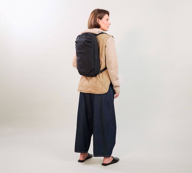 Woman wearing a tan jacket and wide navy trousers, turned away carrying a black backpack