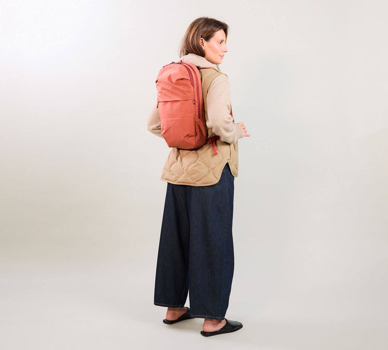 Woman wearing coral backpack and beige jacket, standing and looking over shoulder, navy wide-leg pants and black flats.
