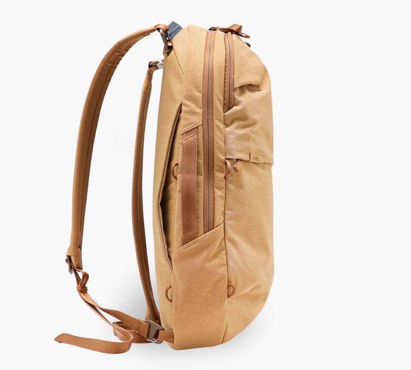 Tan slim backpack side profile showing zippered front pocket, padded shoulder straps, and top grab handle.