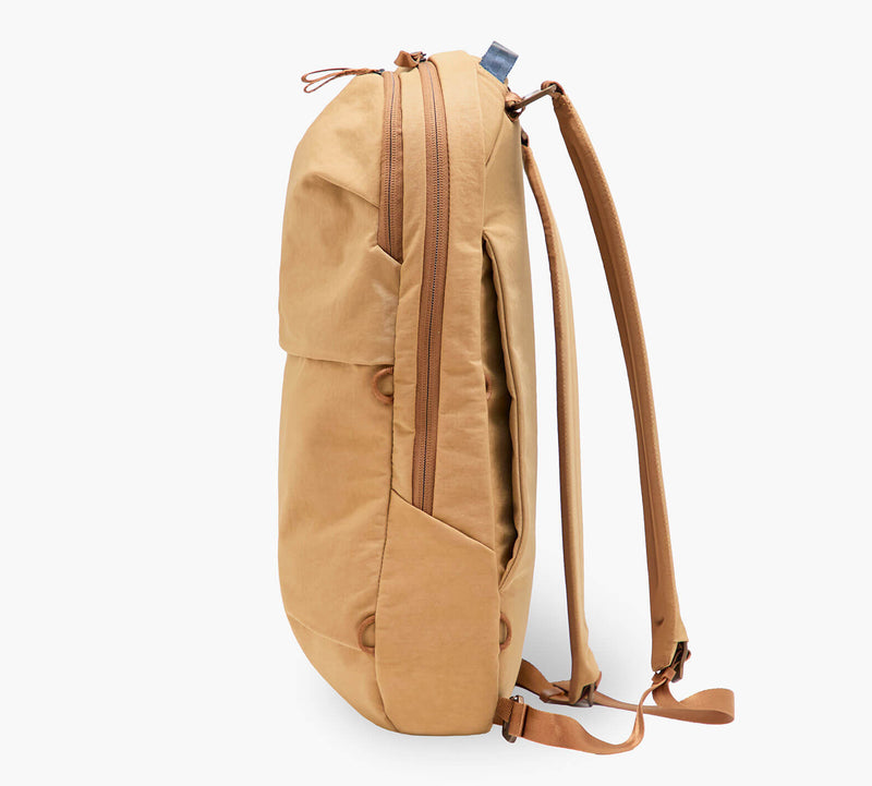 Tan canvas backpack, side view showing zippered main compartment, front pocket, and adjustable shoulder straps.