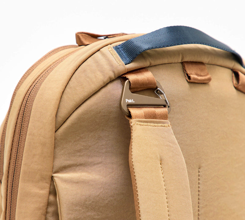 Rear shoulder strap and metal hardware of a tan backpack, showing padded handle and zipper seam.