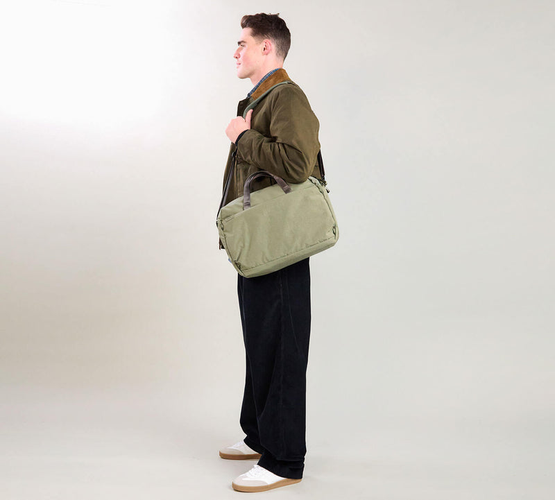 Man in profile carrying an olive-green duffel bag over his shoulder; wearing olive jacket, black trousers, white sneakers.