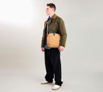 Male model wearing an olive jacket, navy shirt, wide black pants and white sneakers, carrying a tan crossbody bag, standing facing left.