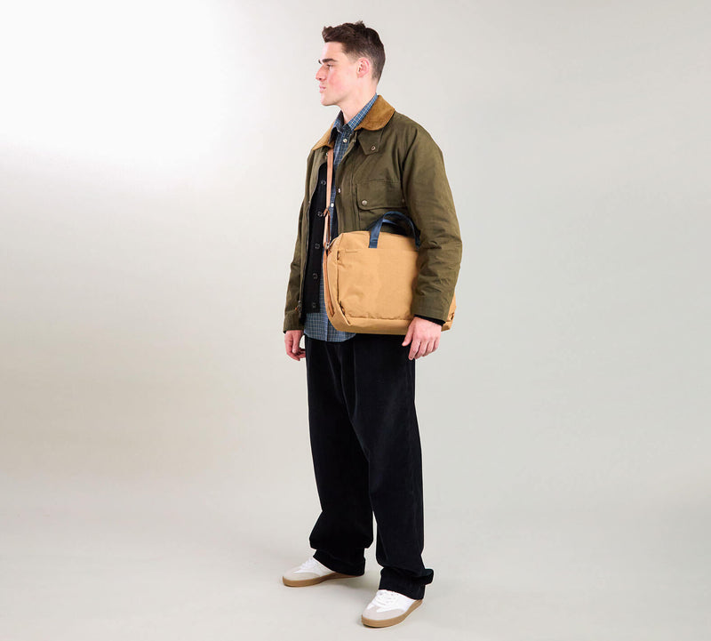 Male model wearing an olive jacket, navy shirt, wide black pants and white sneakers, carrying a tan crossbody bag, standing facing left.