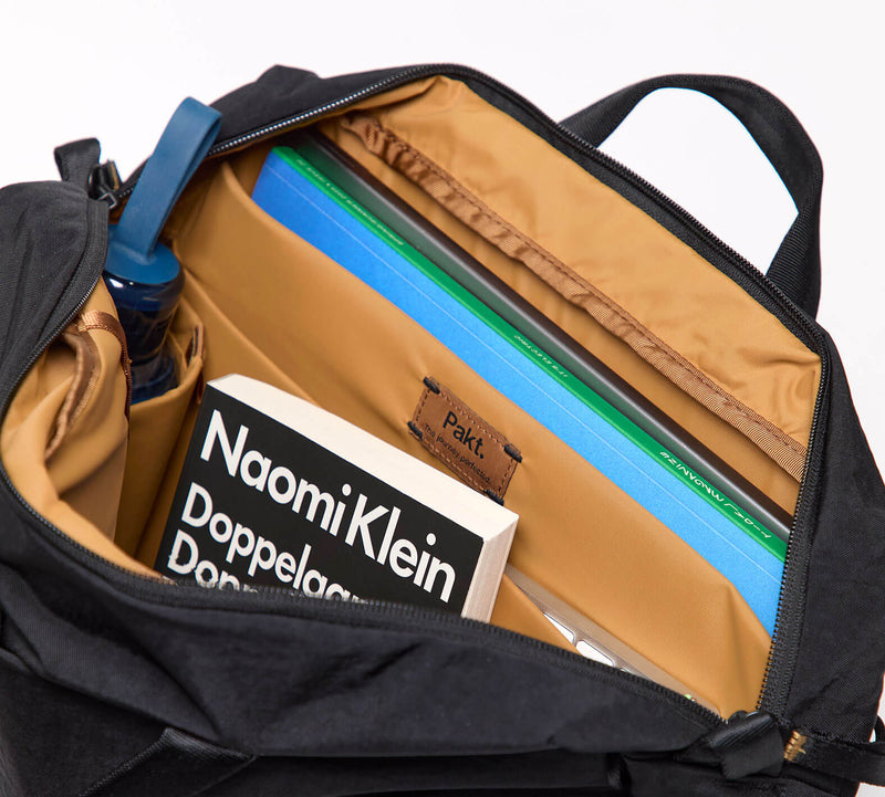 Open shoulder bag interior with tan pockets, a book titled 'Naomi Klein', and leather label 'Pakt.'