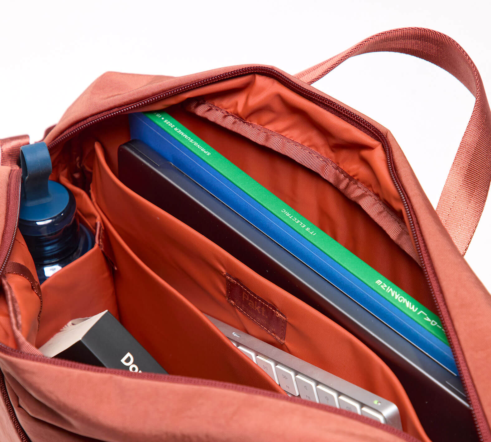 Top-down view of MODE Shoulder Bag 12L in Sumac showcasing open interior with padded laptop sleeve, notebook slots, and water bottle pocket on white background