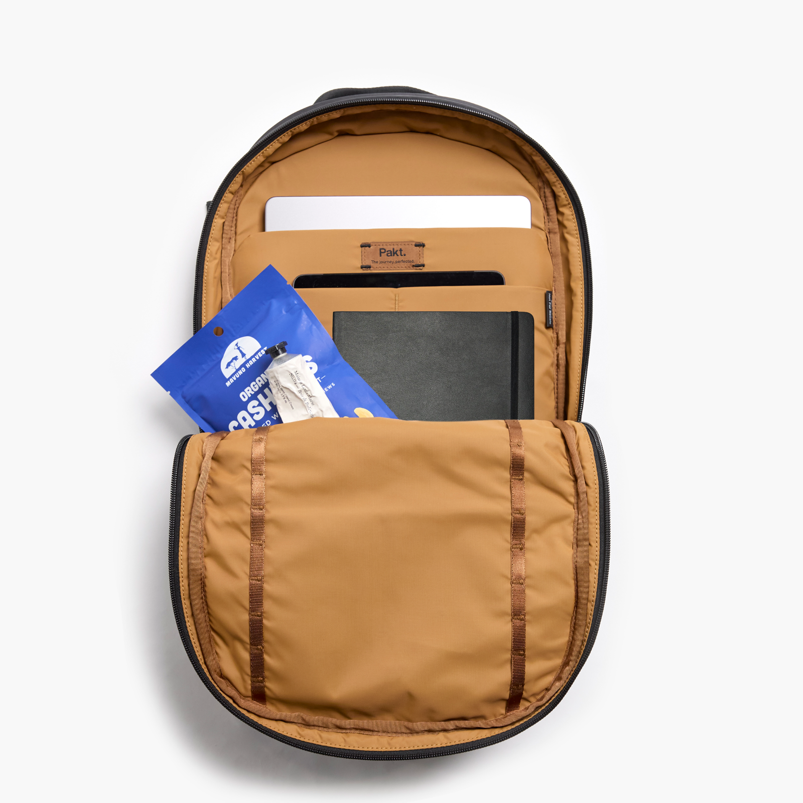 Pakt. backpack interior in mojave with laptop sleeve, notebook, and blue pouch labeled ORGANIC CASHEWS
