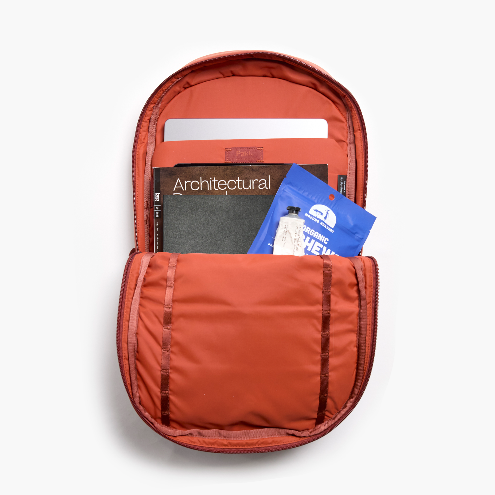 Pakt One Travel Backpack+ open in sumac, laptop sleeve labeled Pakt, book titled Architectural and blue packet ORGANIC CHEW