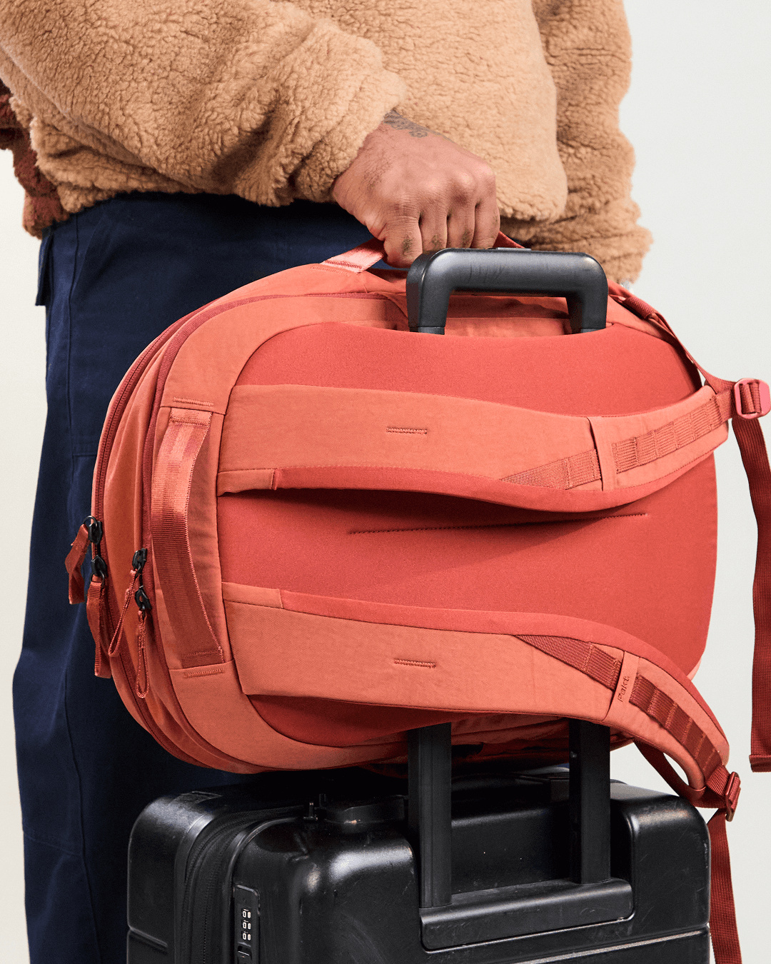 Angled view of MODE Backpack 22L in sumac showing luggage pass-through and padded back panel on a suitcase
