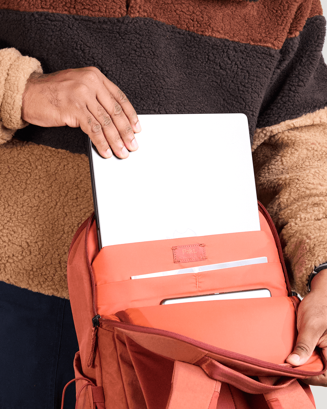 Top-open view of MODE Backpack 22L showing padded 16-inch laptop sleeve and admin pockets in sumac, hands inserting laptop