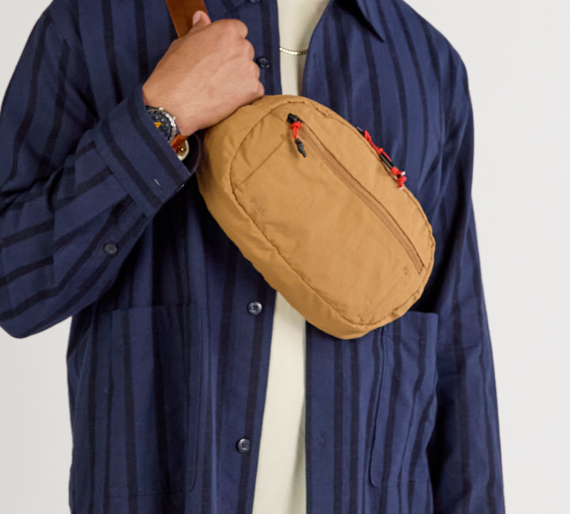 Mojave-colored Stash Sling worn crossbody over navy striped shirt; tan bag with red zipper pulls visible.