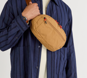 A model wearing a blue shirt with the Stash Sling in Mojave worn over.