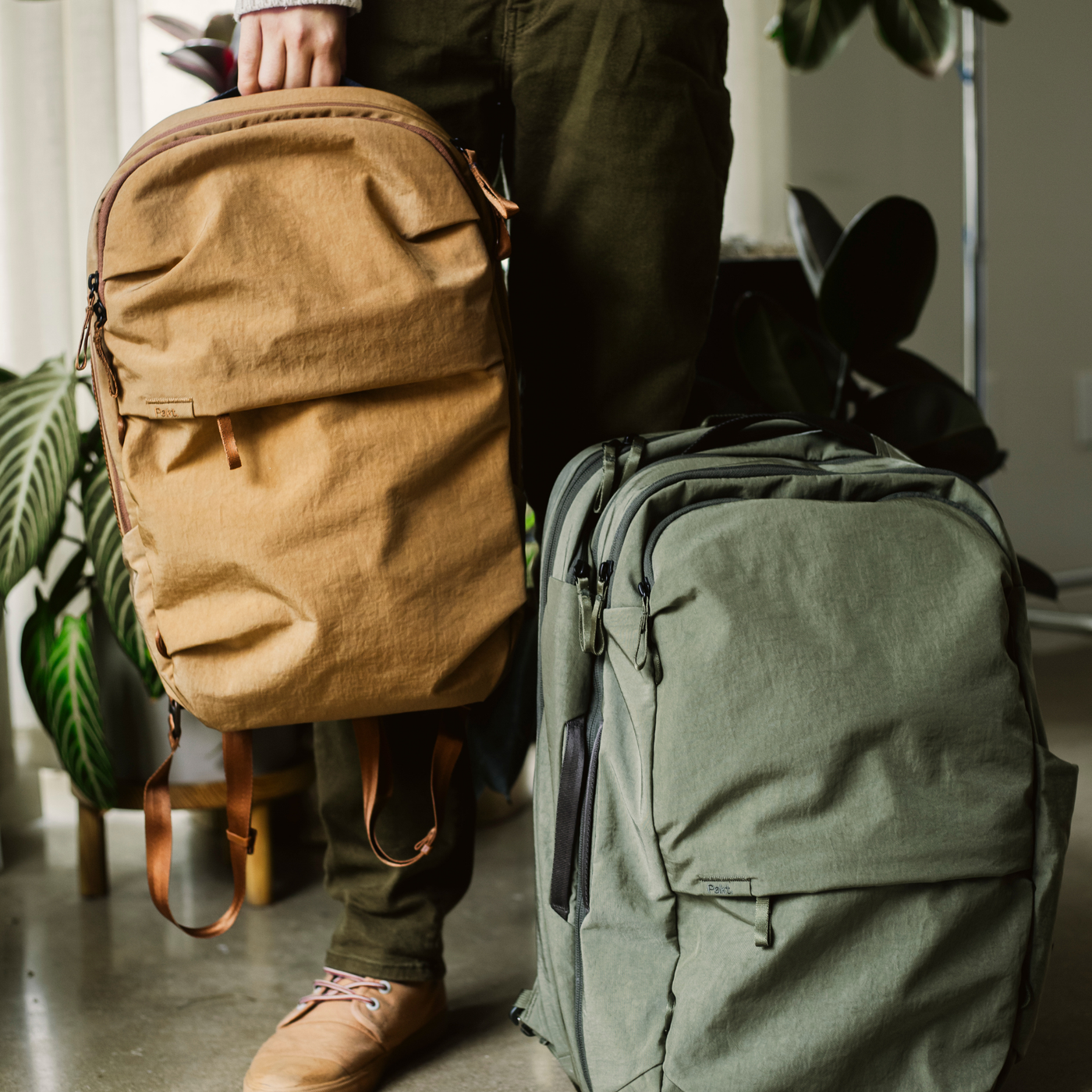 Tan Pakt backpack held by a person next to a green Pakt carry-on backpack on the floor