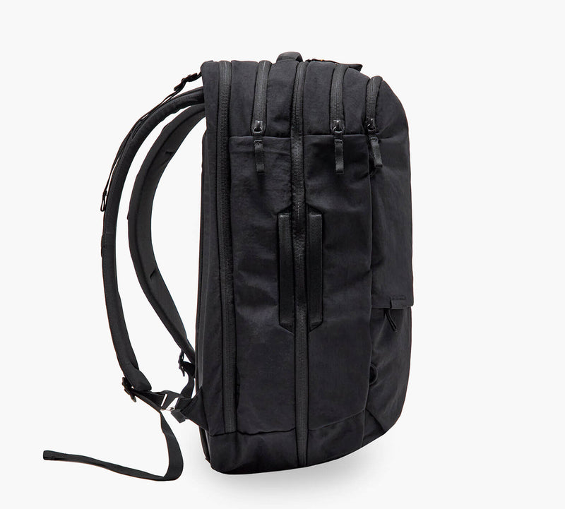 Black backpack, side view showing padded shoulder straps, three zippered compartments, and a side handle.
