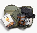 Open olive-green carry-on with brown sandals, packed clothes and a book titled 'The Atlas'.