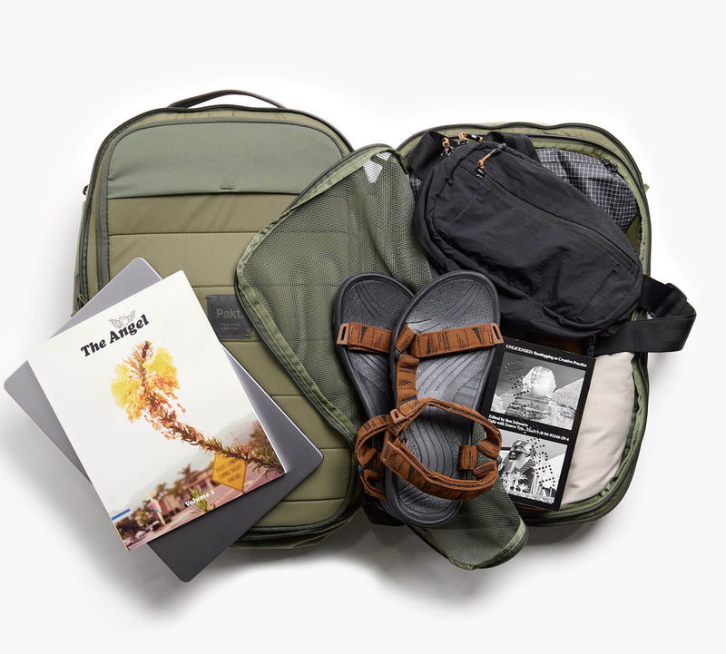 Open olive-green carry-on with brown sandals, packed clothes and a book titled 'The Atlas'.