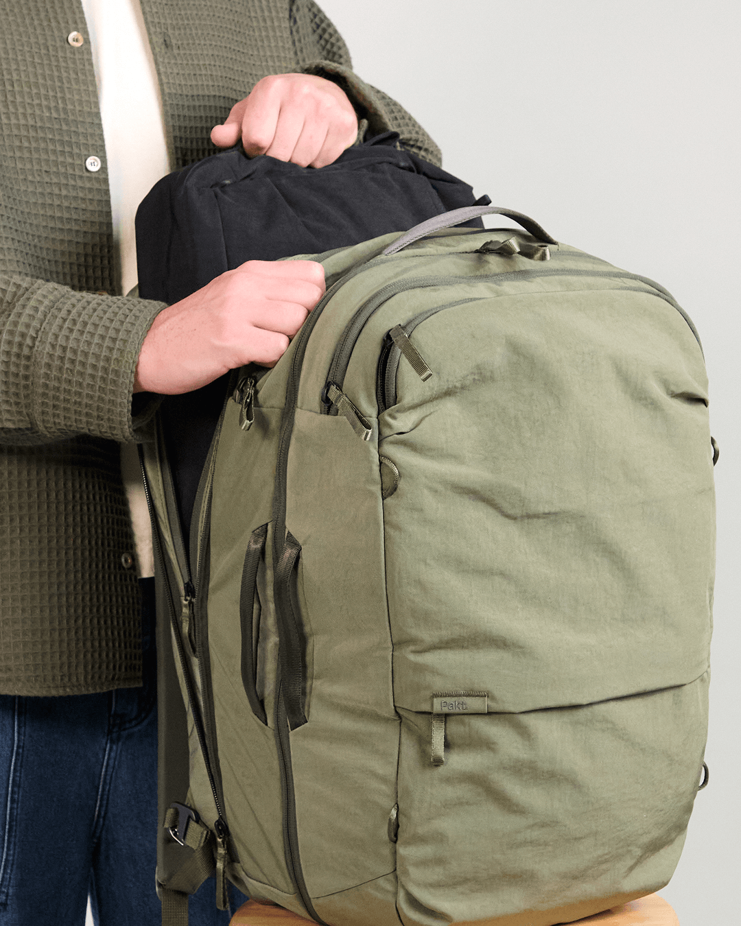 Angled front view of Pakt One backpack in cypress showing open compartment with packing cube and handles on gray background