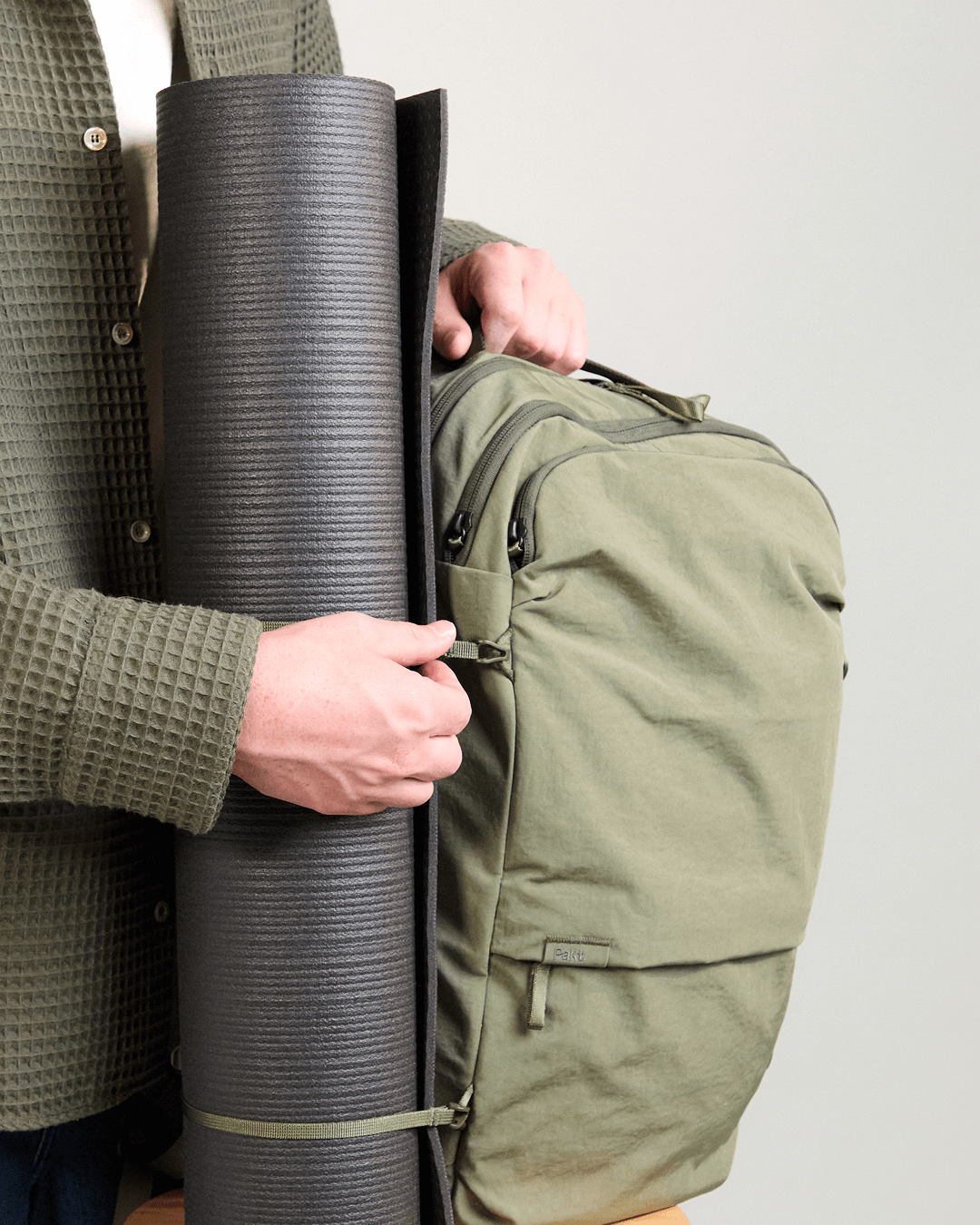 Side view of Pakt One Travel Backpack in cypress showcasing stowable straps securing a rolled yoga mat and front zip pocket on a light gray background