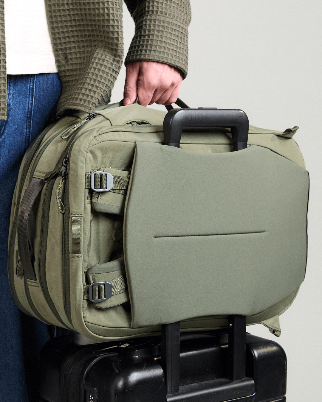Side view of Pakt One Travel Backpack in cypress showing luggage pass-through sleeve on rolling suitcase and stowed hip belt