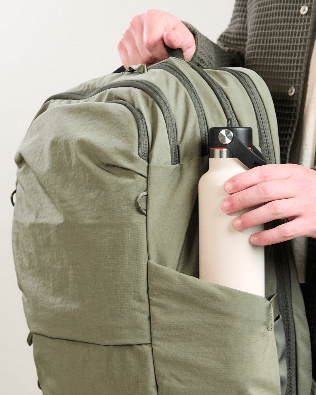 Hand placing a cream insulated water bottle into the side pocket of an olive-green backpack