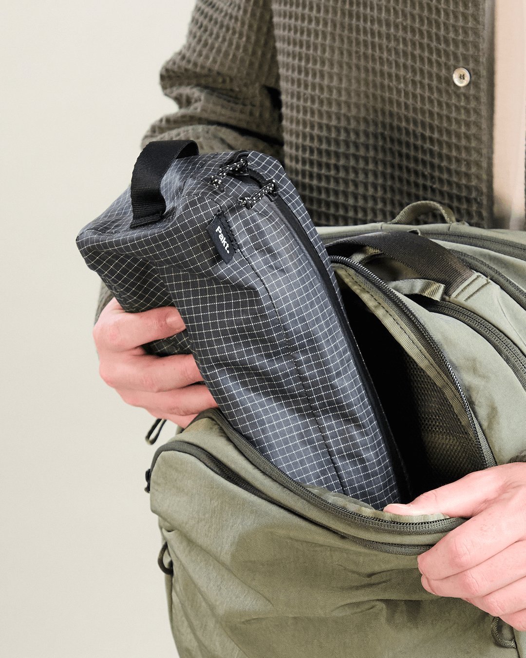 Close-up of Pakt One Travel Backpack in cypress showing the front compartment open as a hand inserts a gray grid-pattern accessory pouch