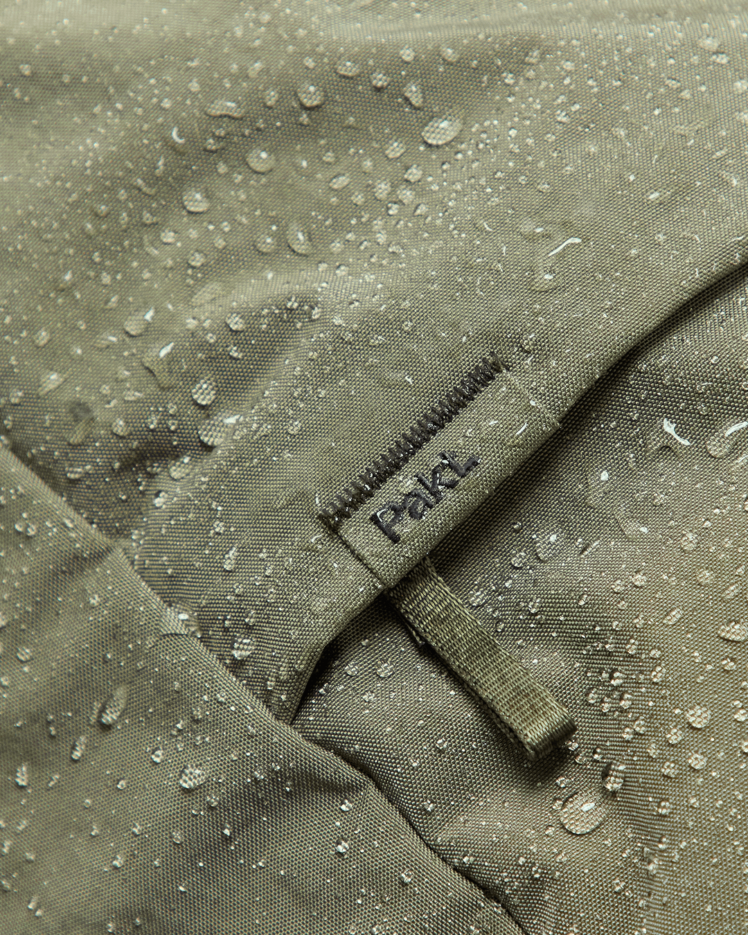 Close-up of Pakt One Travel Backpack exterior in cypress showing water beading on the fabric and a sewn Pakt tag