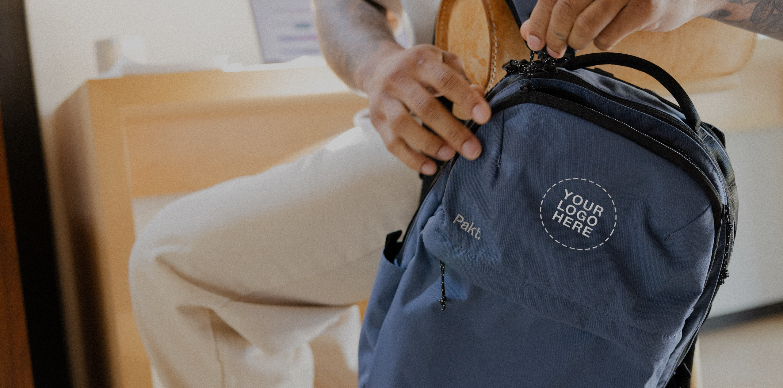 Navy backpack with circular patch reading "YOUR LOGO HERE" and small Pakt. label; hands adjusting the top zipper.