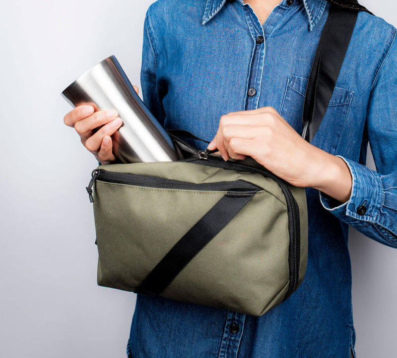 Person in denim shirt placing stainless-steel travel mug into olive-green crossbody bag