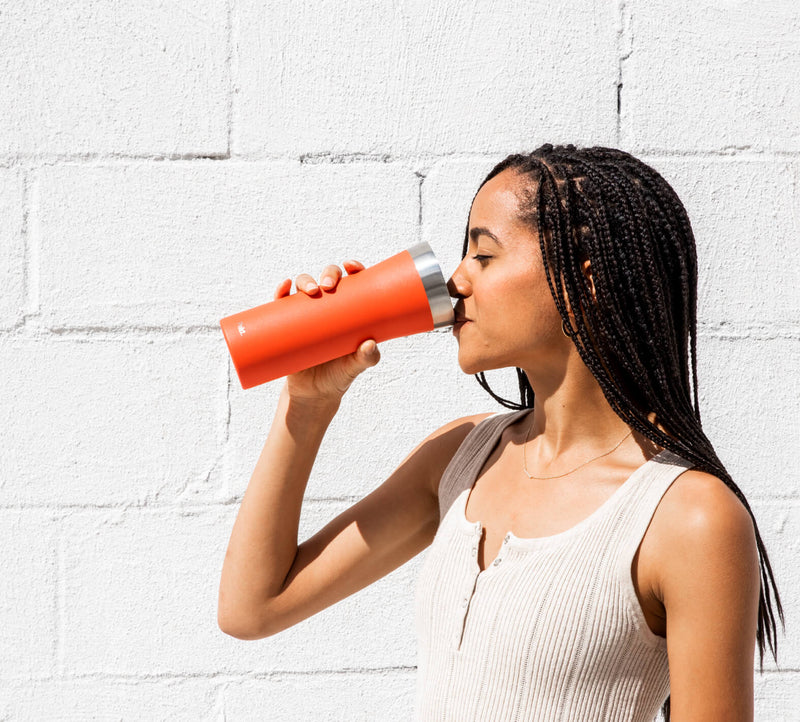 Young woman with braided hair sipping from an orange travel tumbler beside a white brick wall